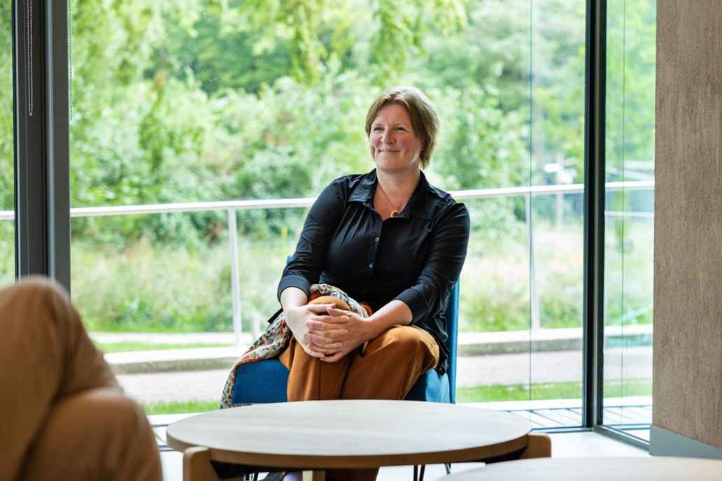 Liesbeth Soer, directeur Catalytic Investments. Liesbeth zit op een blauw stoeltje voor een groot raam. Ze draagt een zwarte bloes en een cognac kleurige broek.